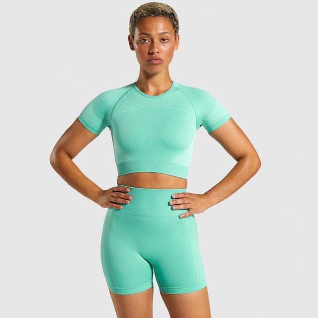 Girl wearing 2 piece yoga set. Bottoms are long shorts. Top has a high rounded neck line and short sleeves. Color is green 